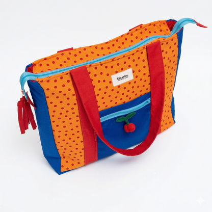 Bolsa Tote Cerezas — Encantos con diseño en naranja y azul, detalles en rojo, cierre celeste y pequeño adorno de cerezas tejido en el bolsillo frontal.