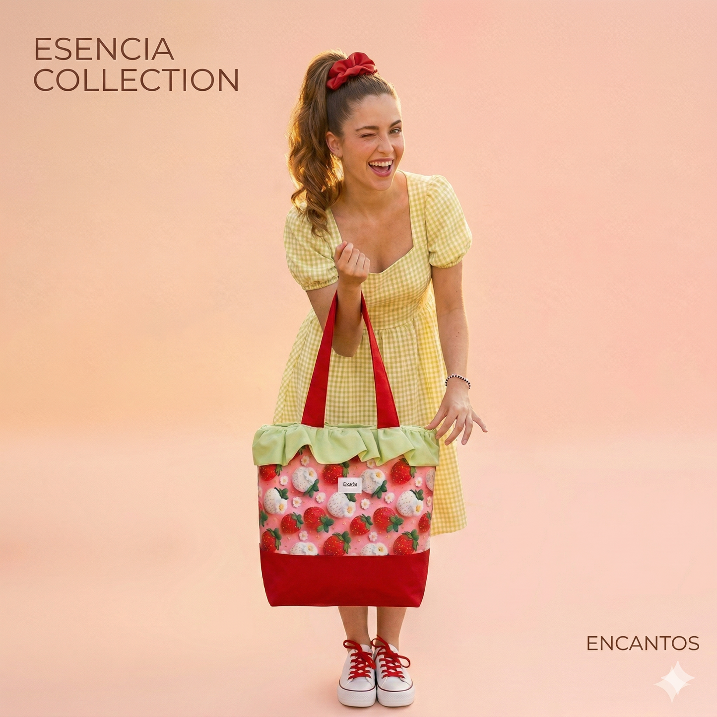 Mujer sonriente con vestido amarillo sostiene un Bolso Tote Fresas con Volante Verde de diseño fresco con fresas y detalle de volante sobre fondo rosa suave.